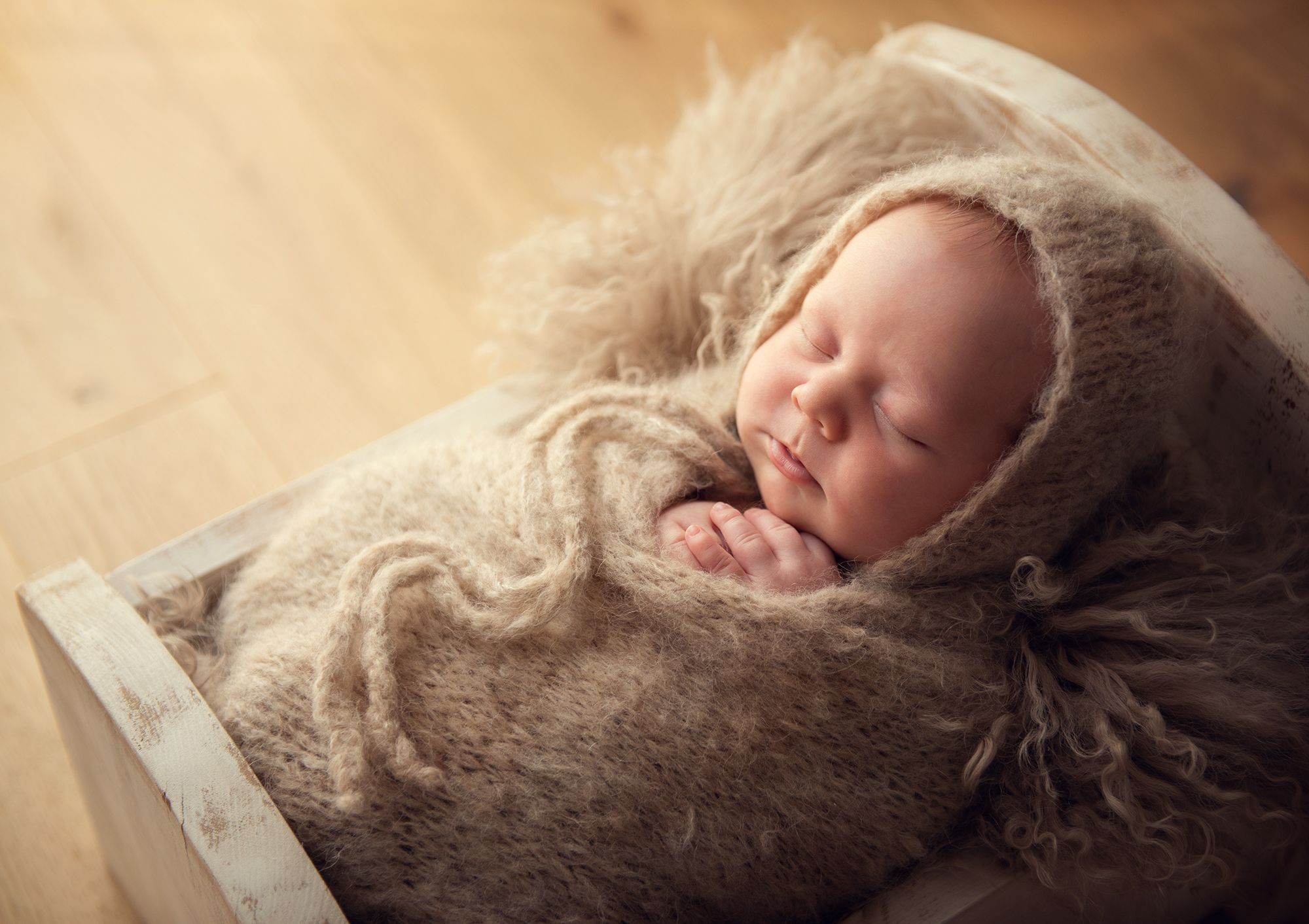 A sleeping newborn baby wrapped in brown textured blanket lies peacefully in soft natural lighting.