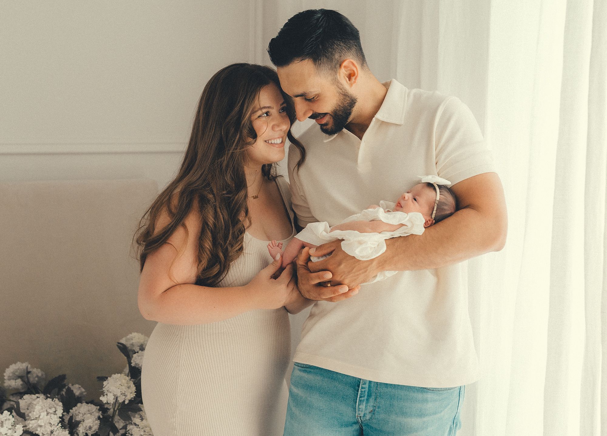 A couple tenderly holds their newborn baby, smiling softly in a bright, elegant room with floral decor.