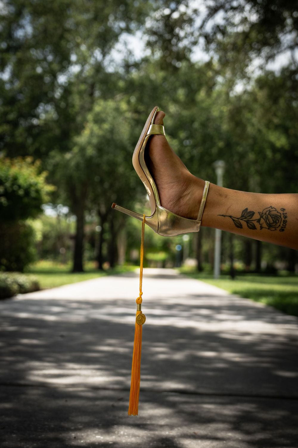 A tattooed foot in a gold stiletto heel holds a graduation tassel outdoors on a tree-lined path.