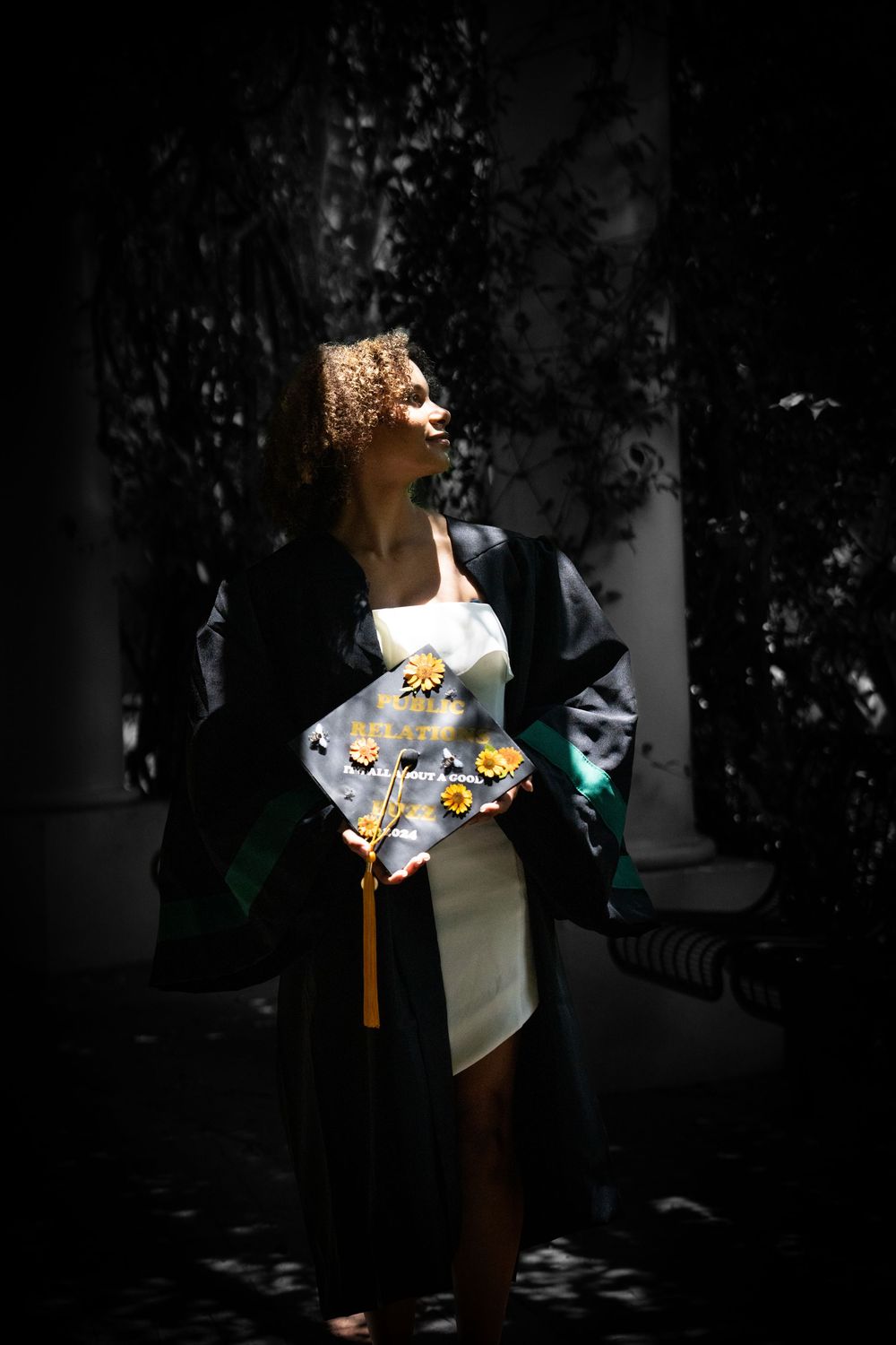 A young woman in graduation regalia holds a decorated cap with yellow flowers, standing in dramatic light and shadow.