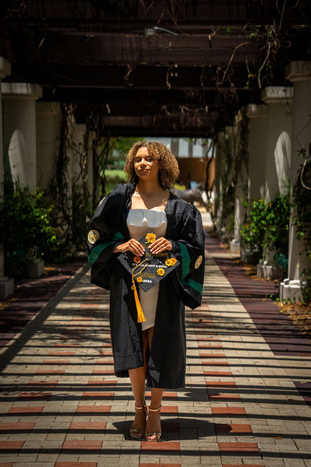 Young woman in graduation gown walks under pergola, holding cap with yellow tassel on sunny brick pathway.