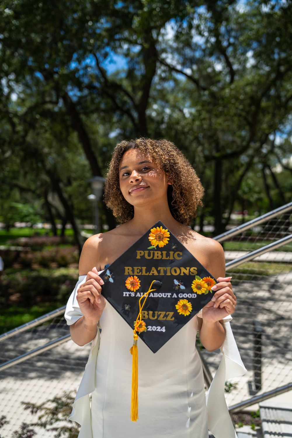 Young woman holding a decorated graduation cap reading Public Relations It's All About a Good Buzz 2024 with sunflowers.