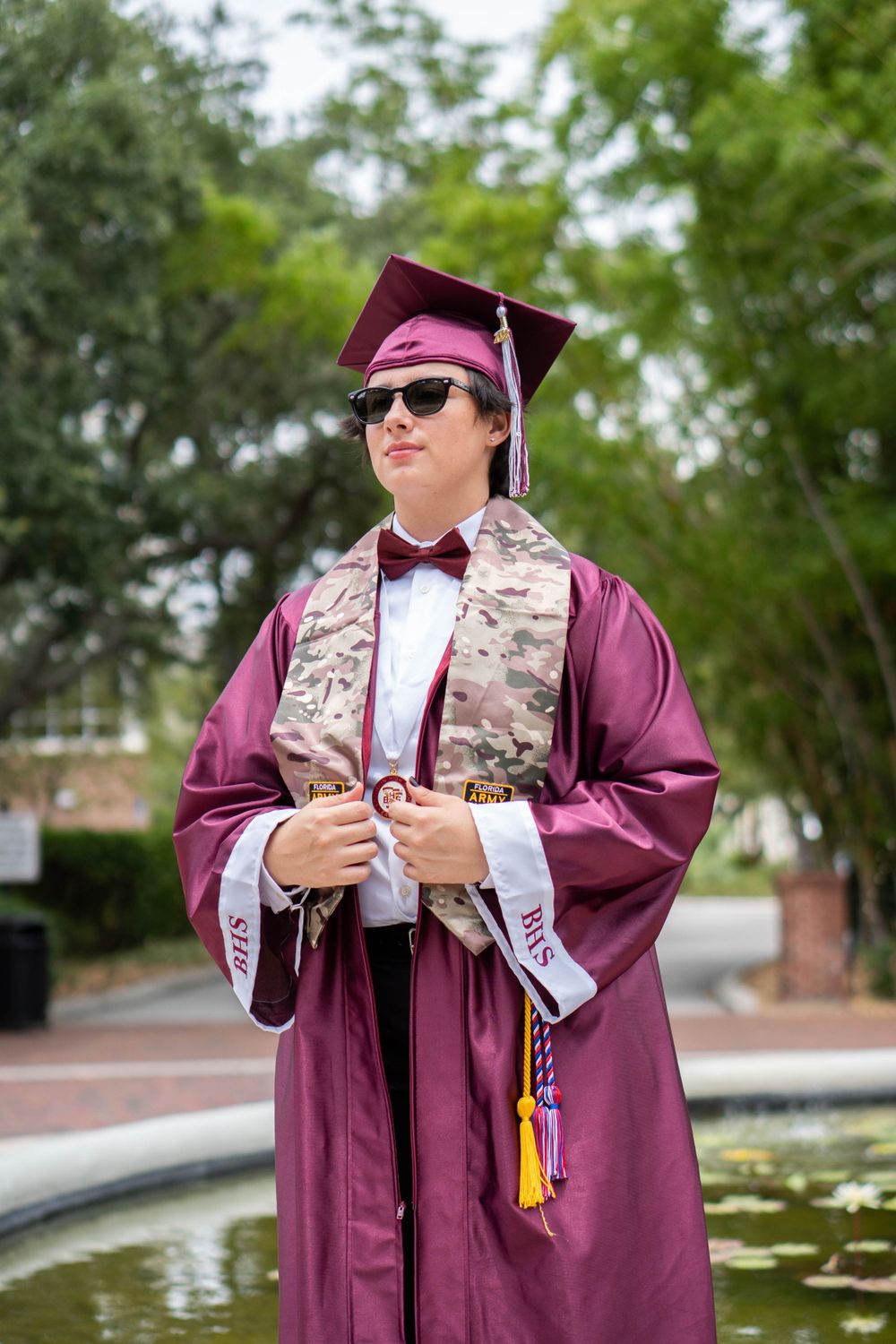Male graduate in maroon cap and gown with sunglasses and bow tie posing near an outdoor fountain in Tampa.