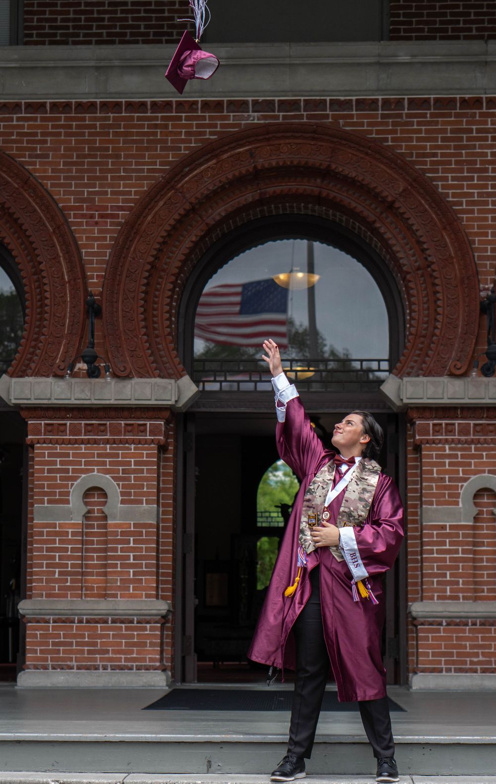 Graduate in maroon gown tosses cap in front of historic brick building with American flag in Tampa.