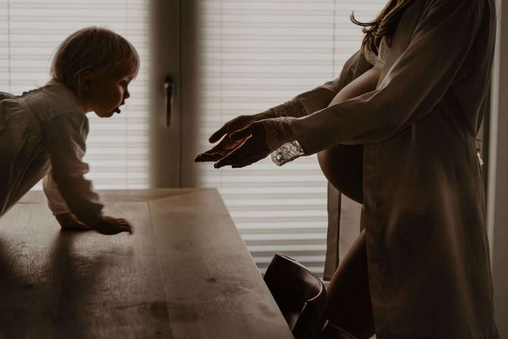 Fotografia artística de gravidez em casa, captando a ternura entre mãe e filha num momento íntimo e cheio de luz natural suave.