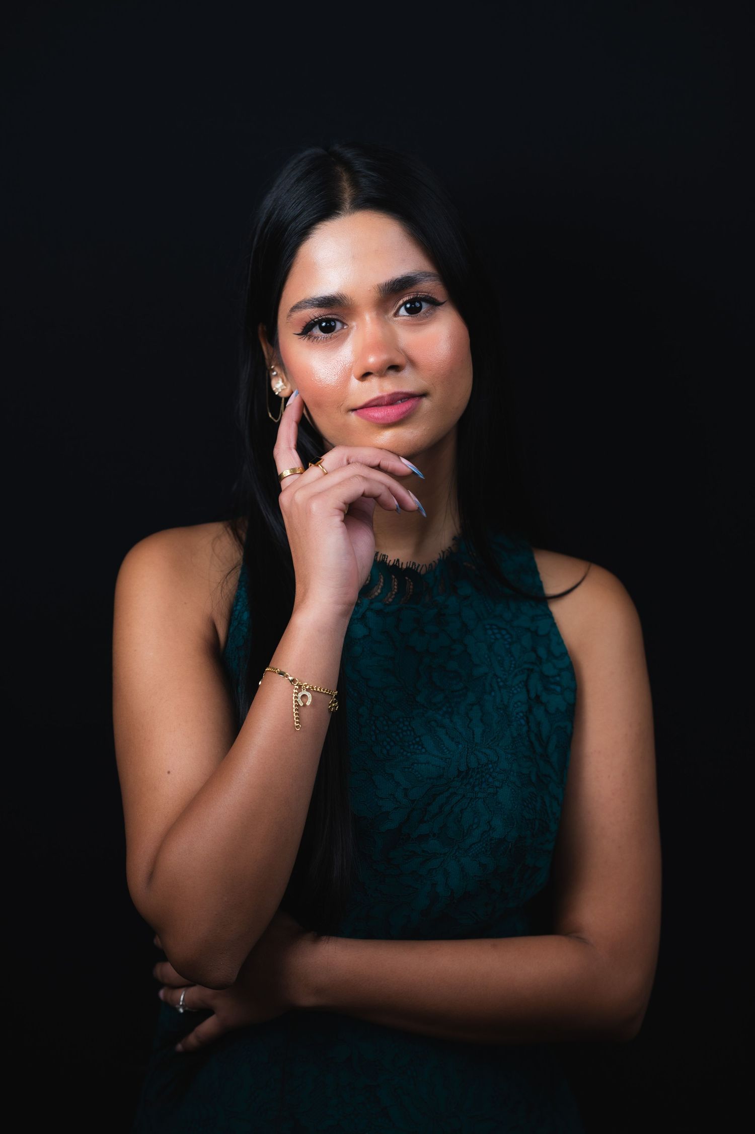 Young woman in teal dress poses thoughtfully with hand on chin against a dark studio background.