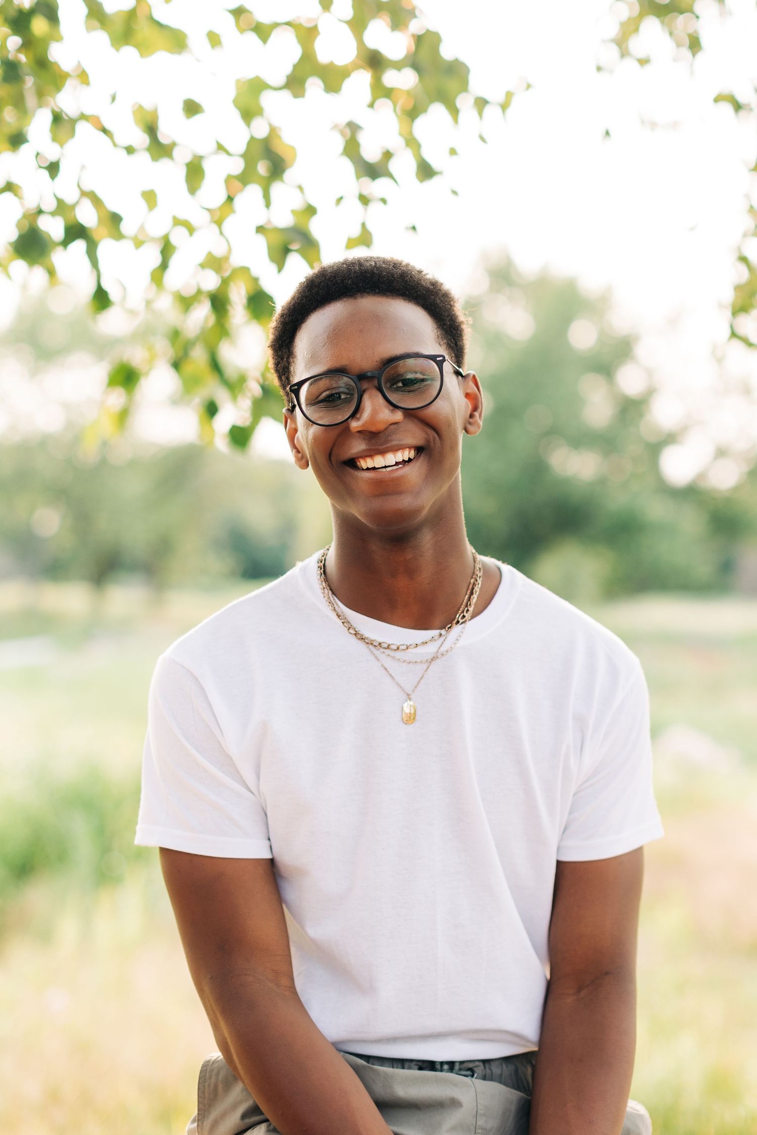 Portrait of someone in a white t-shirt wearing glasses and jewelry against a natural outdoor background.