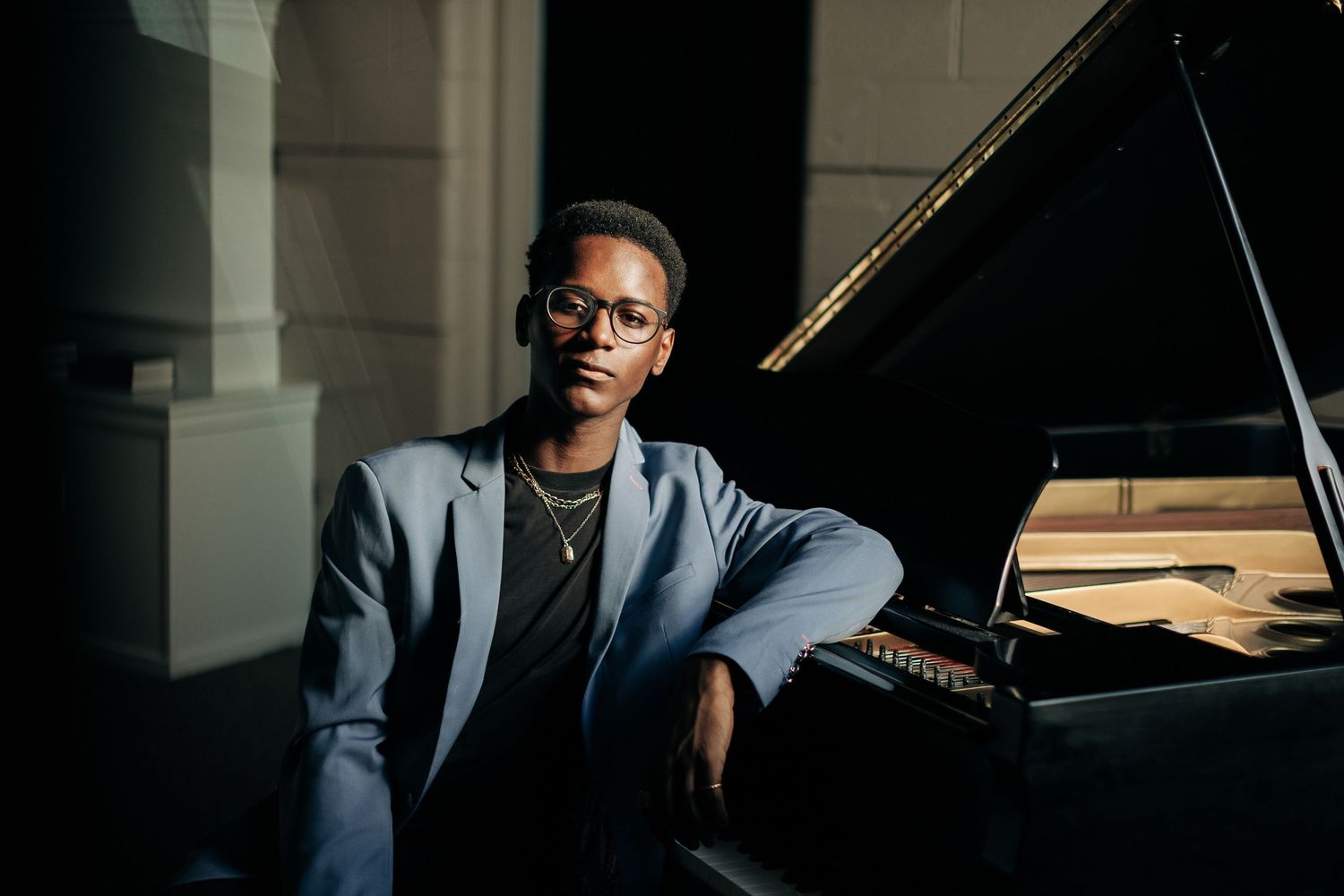 Musician in grey suit jacket sitting at grand piano in dramatic lighting.