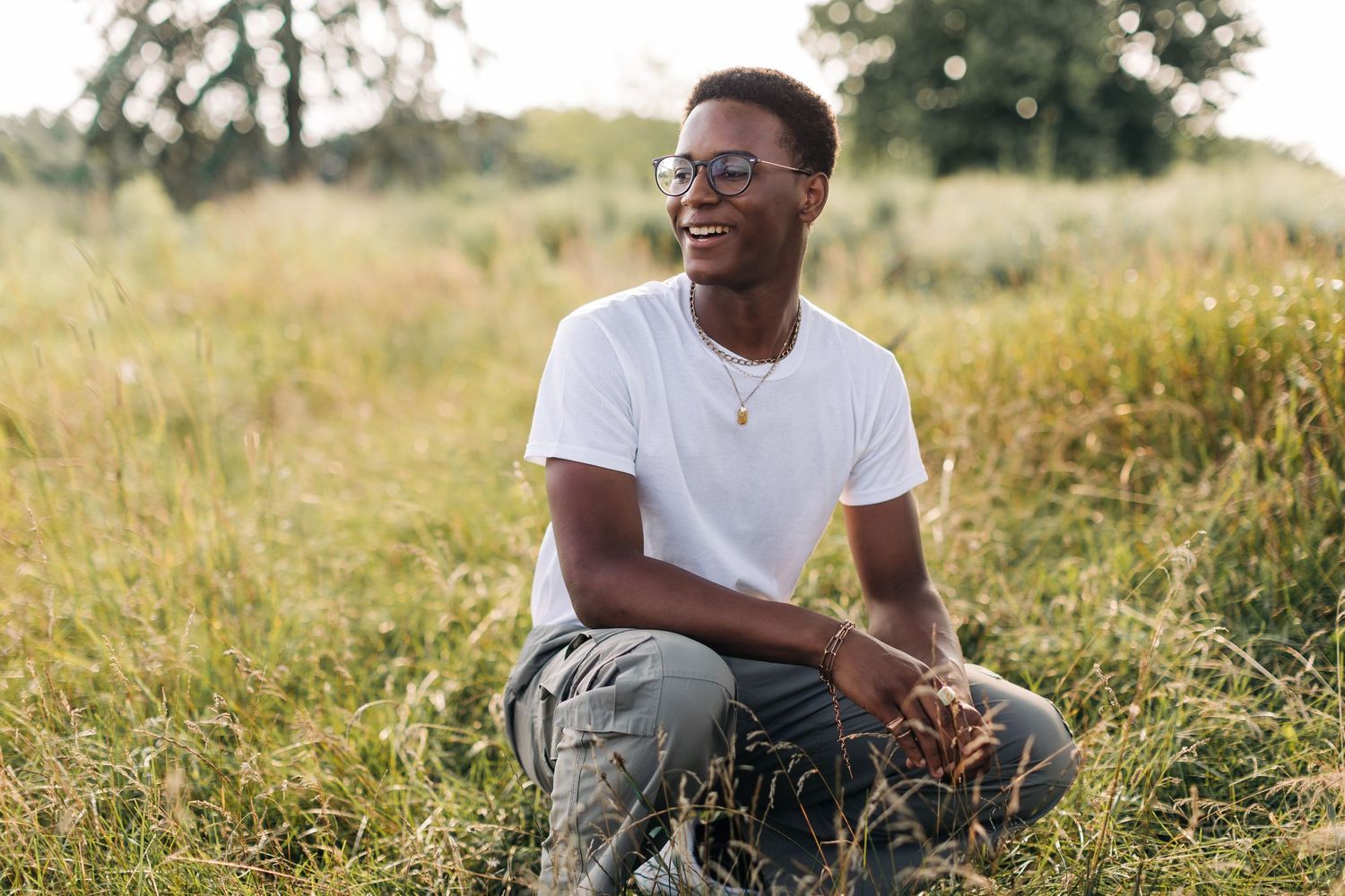 Person wearing glasses and white t-shirt sits in tall grass during golden hour.
