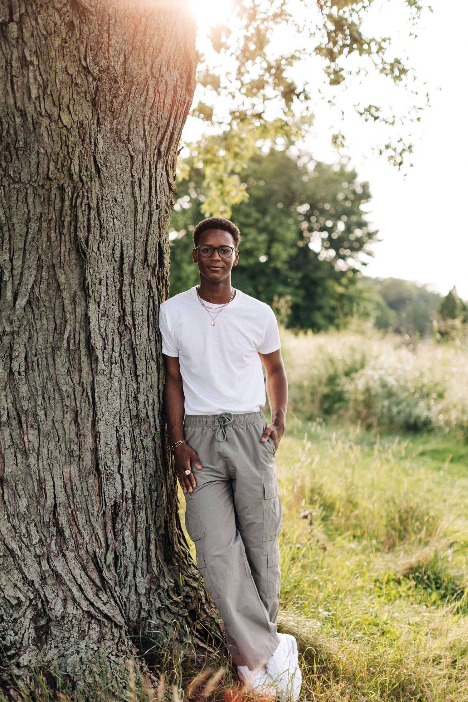 Person in casual attire leaning against large tree trunk in sunlit field.