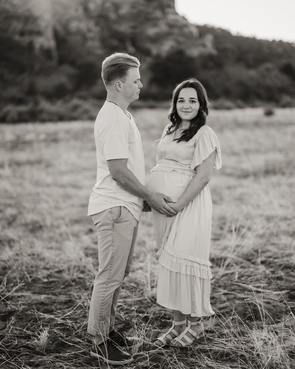 Black and white maternity portrait with couple in light colored clothing against mountainous backdrop.