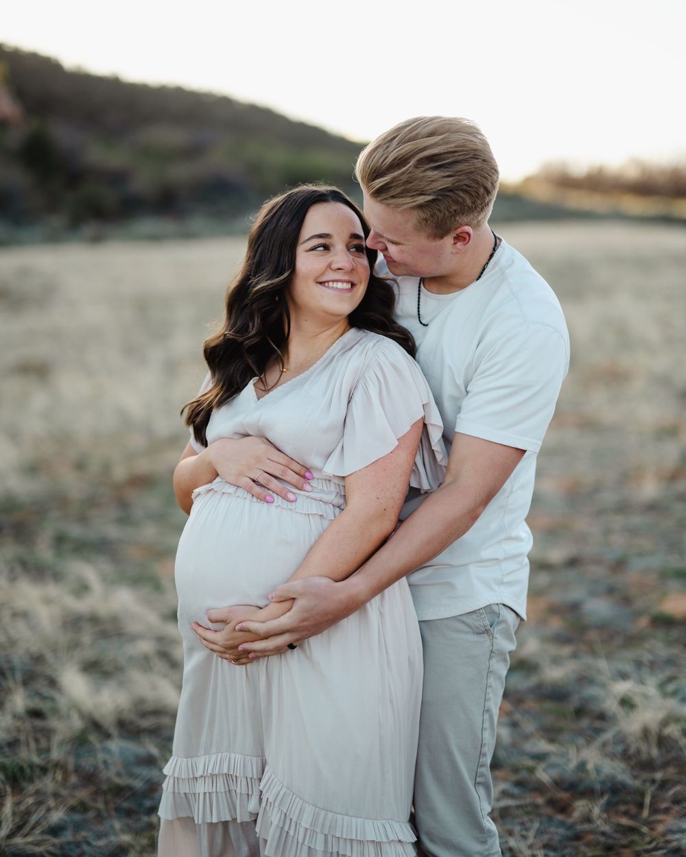 Intimate maternity photo with couple in light clothing against mountain landscape.