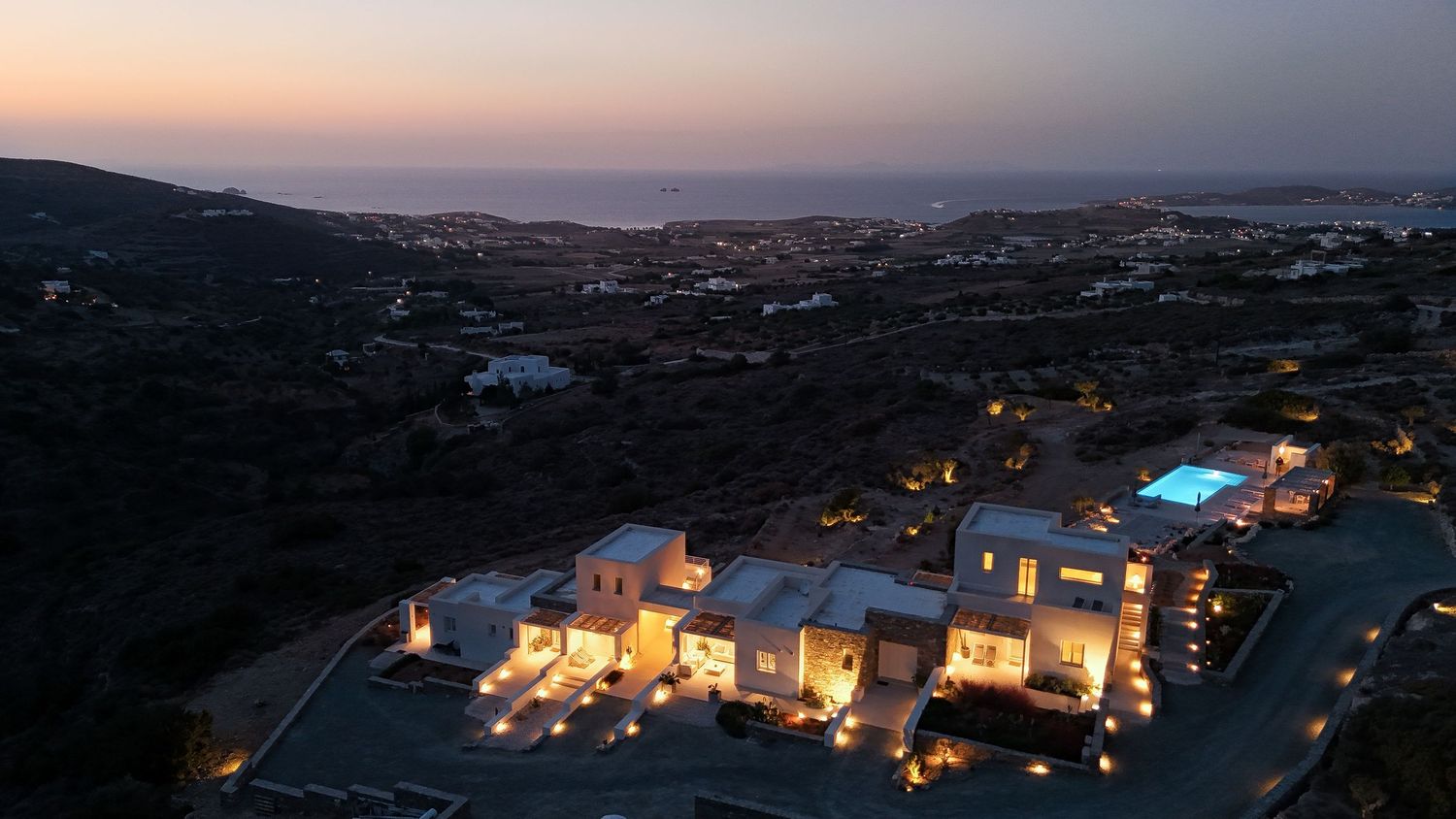 Aerial view of luxury resort villas with swimming pools glowing at sunset against coastal landscape.