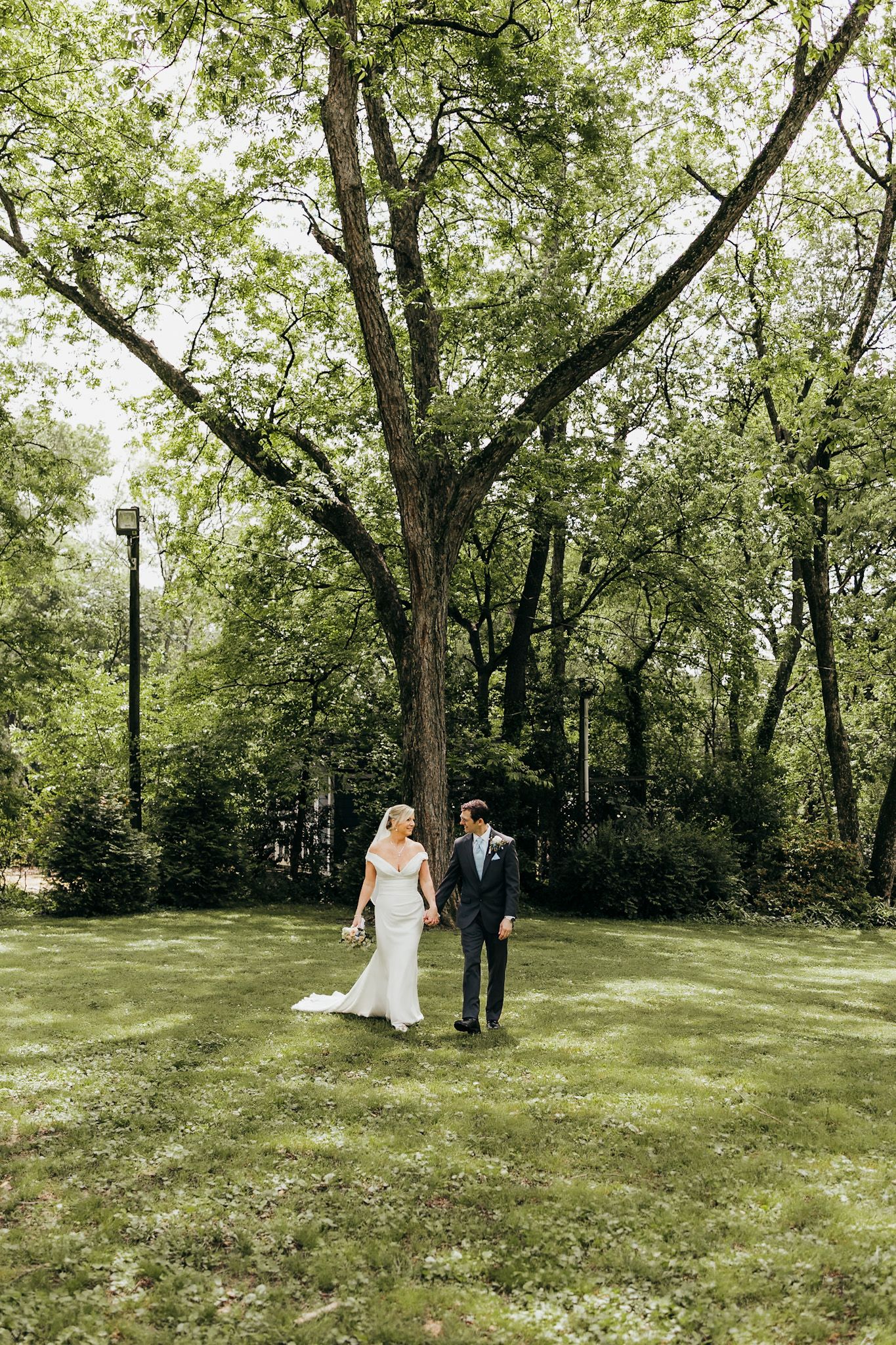 bride and groom holding hands walking in front of a big oak tree. Very green and spring