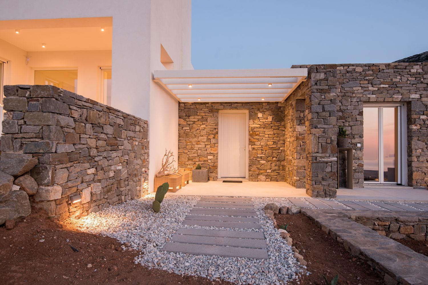 Modern villa entrance with stone walls and white gravel pathway illuminated by warm lighting.