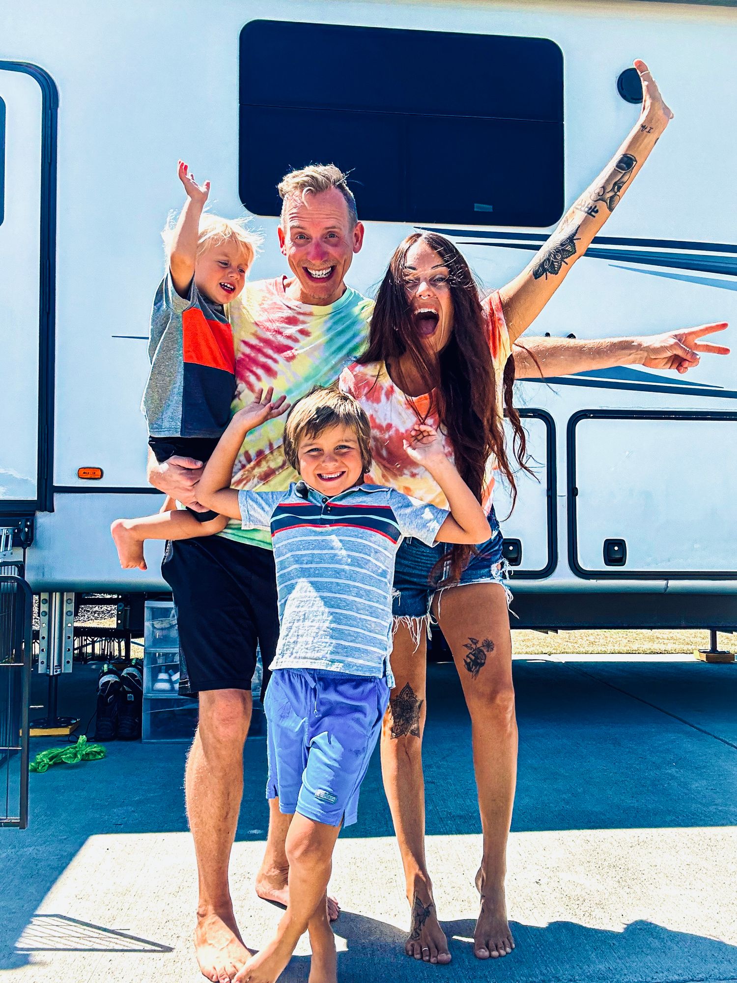 Happy family of four posing energetically in front of a large white RV on a sunny day.