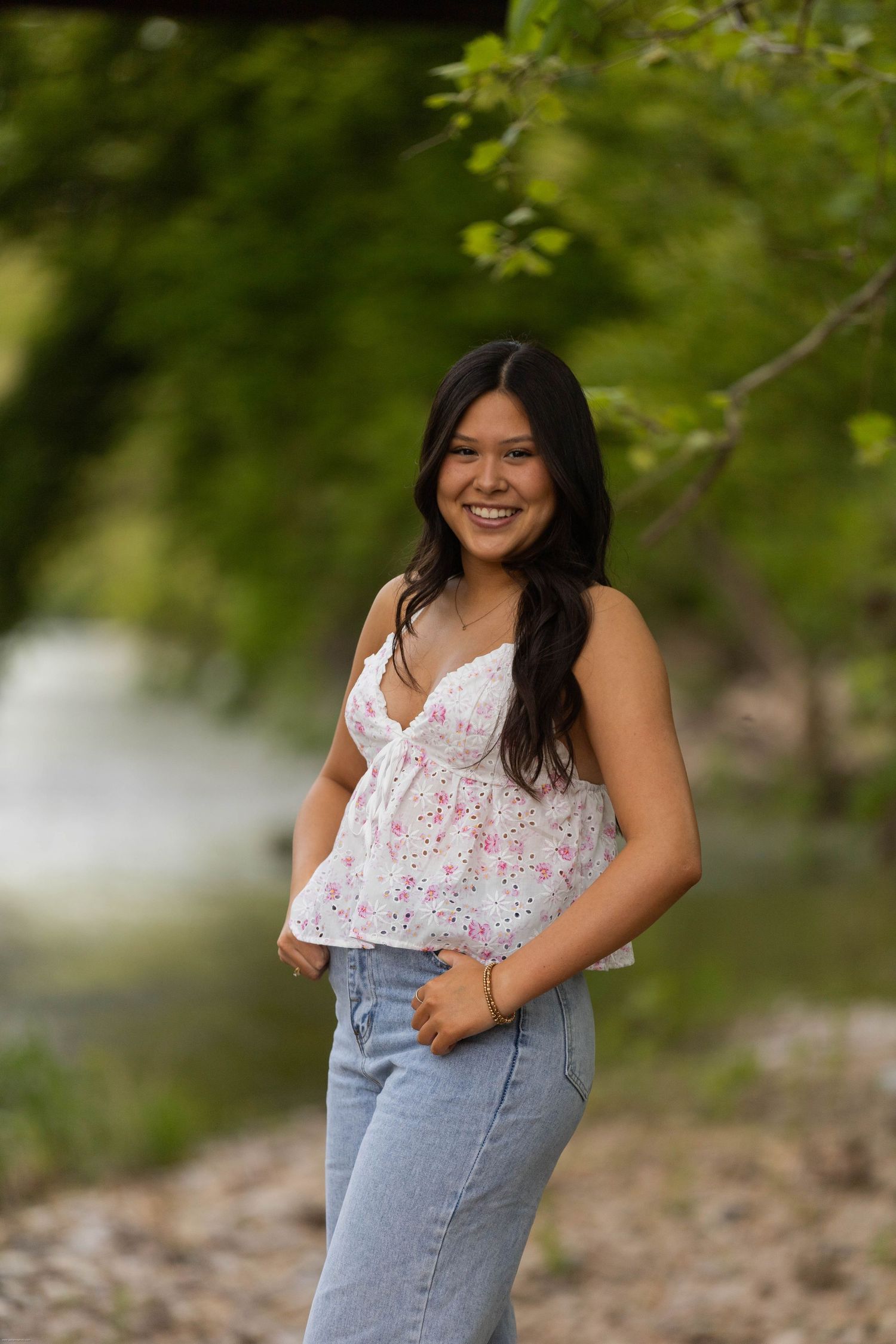 Young person in floral top and jeans poses outdoors against natural foliage background.