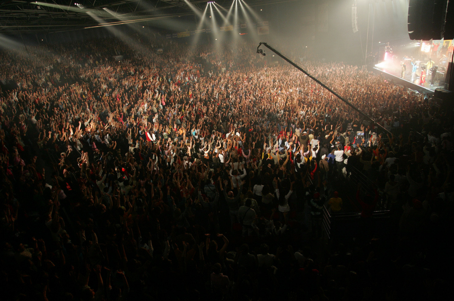 A large crowd of concert attendees fills a dark arena with stage lights and rigging visible overhead.