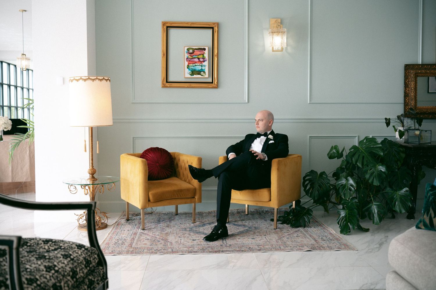 A well-dressed groom sits in a yellow armchair in an elegant room with mint green walls and potted plants.