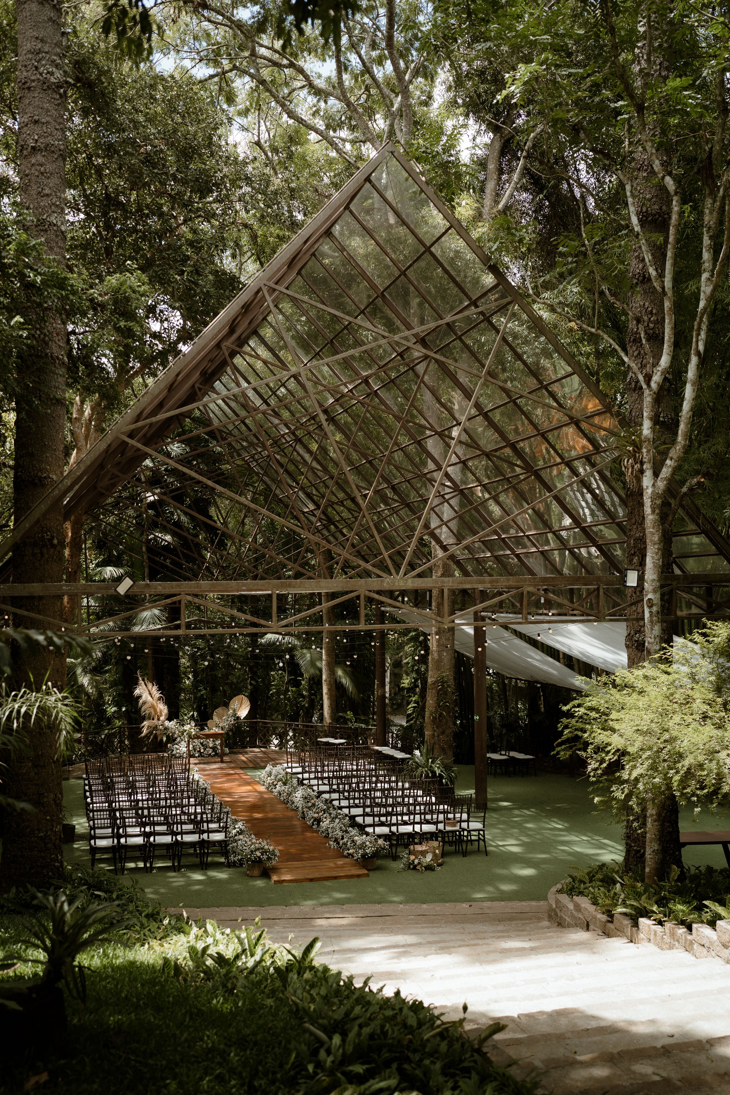 Open-air chapel structure with rows of ceremony chairs and wooden beams framed by lush forest.