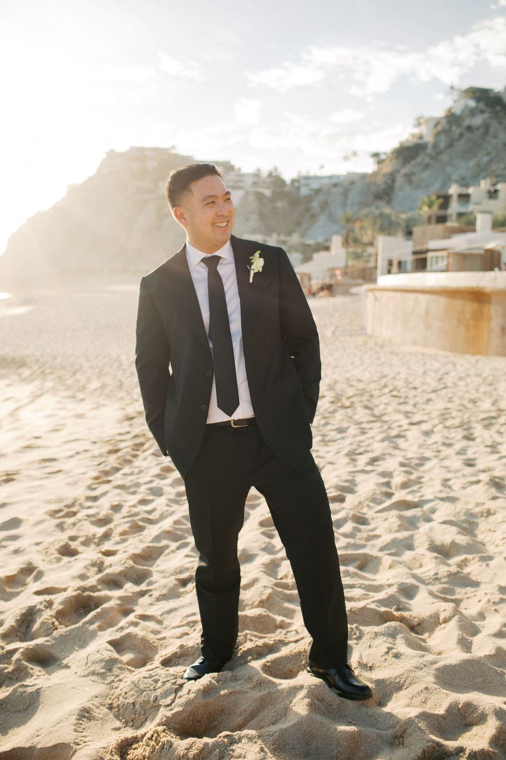 Person in black suit stands on sandy beach with mountains and buildings in background.