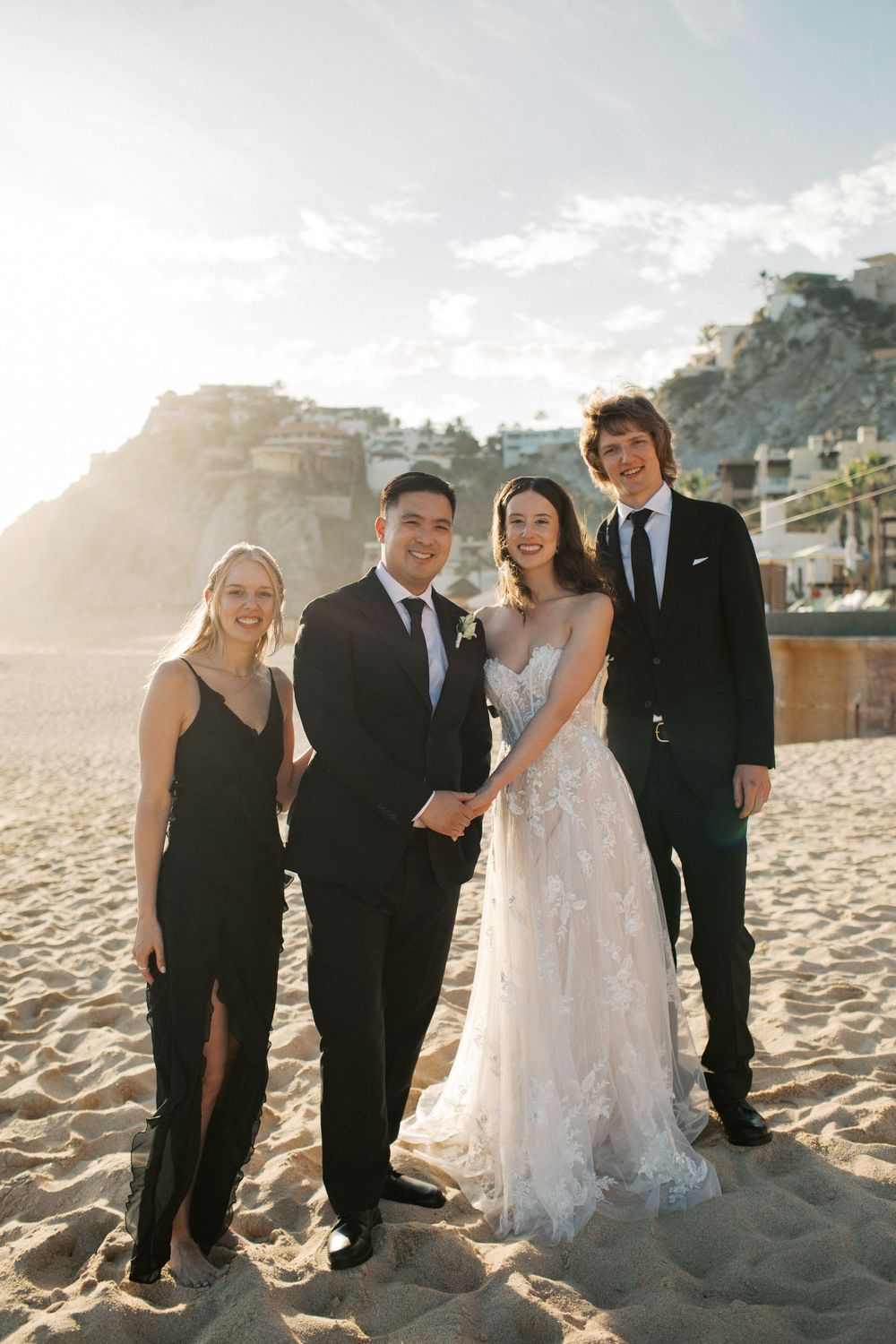 A wedding party poses together on a sunny beach with cliffs and ocean views in the background.