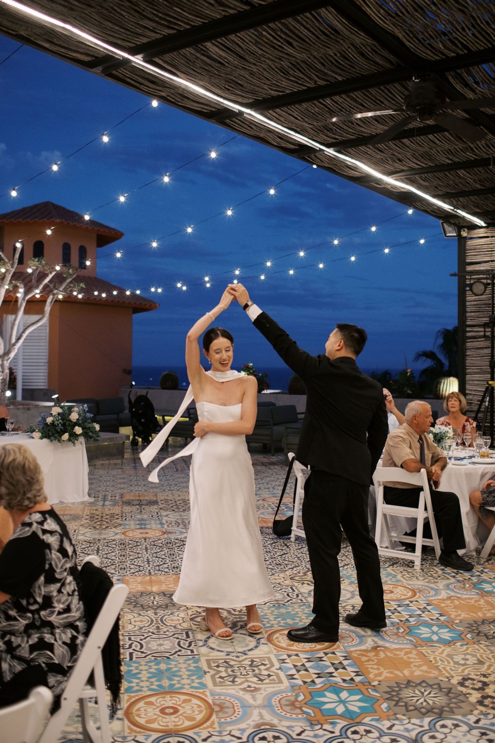 A couple dances under string lights at an outdoor reception with decorative tile flooring.