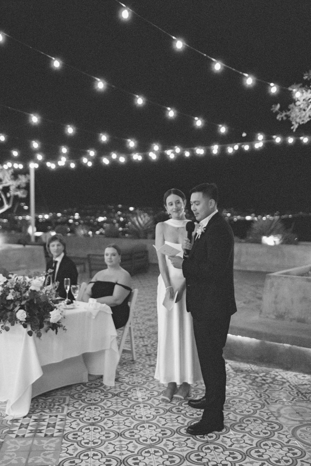 Romantic outdoor wedding reception with string lights and patterned tile floor showing couples dancing at night.