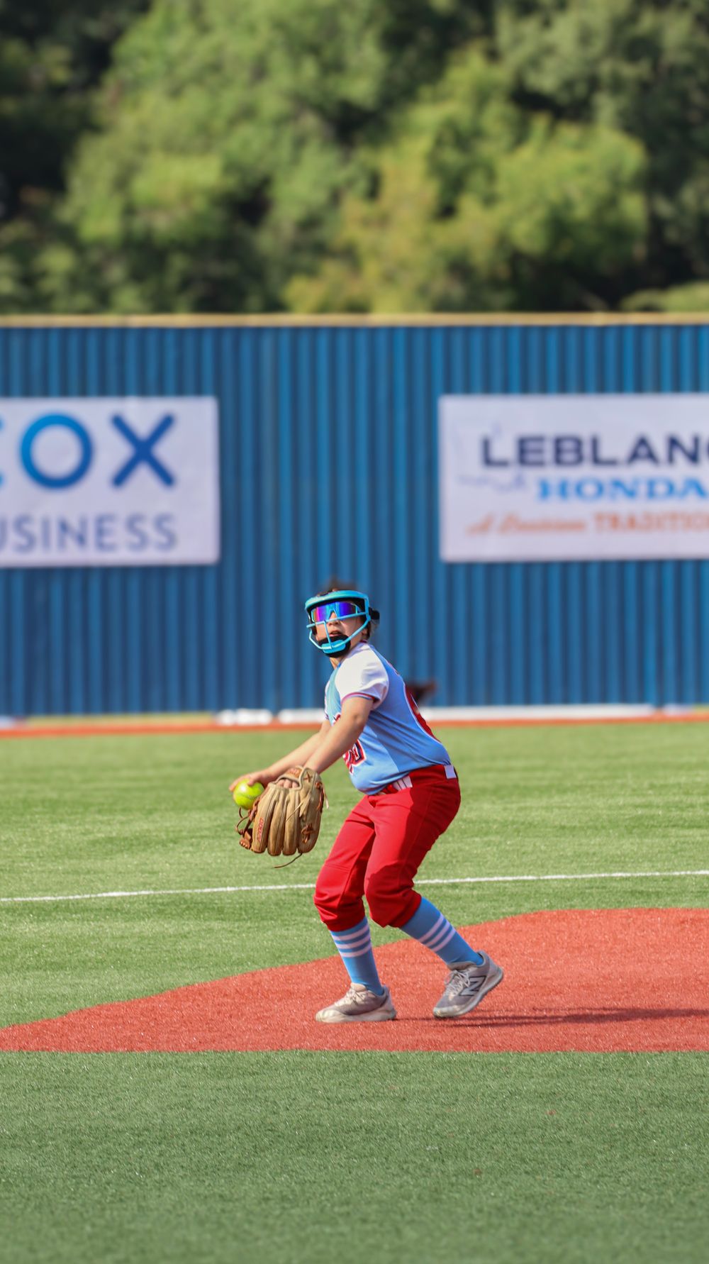 Softball player in red pants and light blue uniform stands ready in fielding position on artificial turf field.