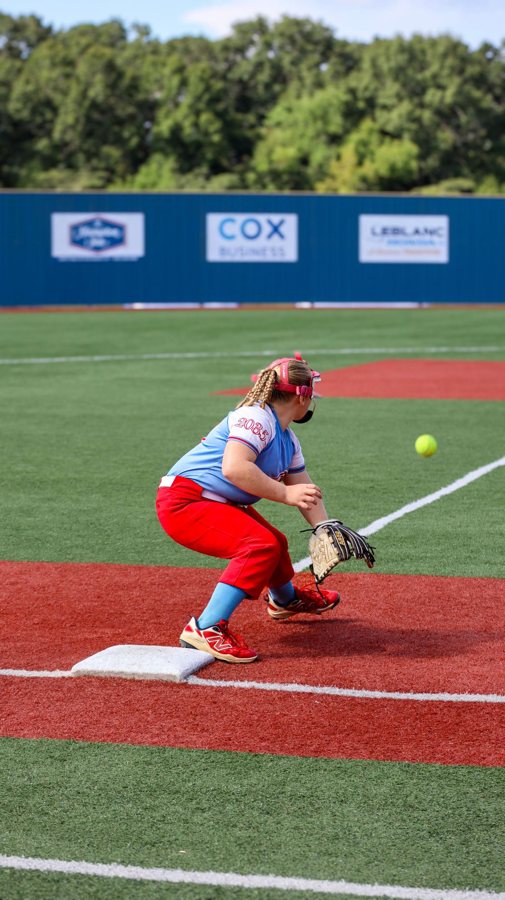 Softball player fielding position at first base with yellow ball approaching.