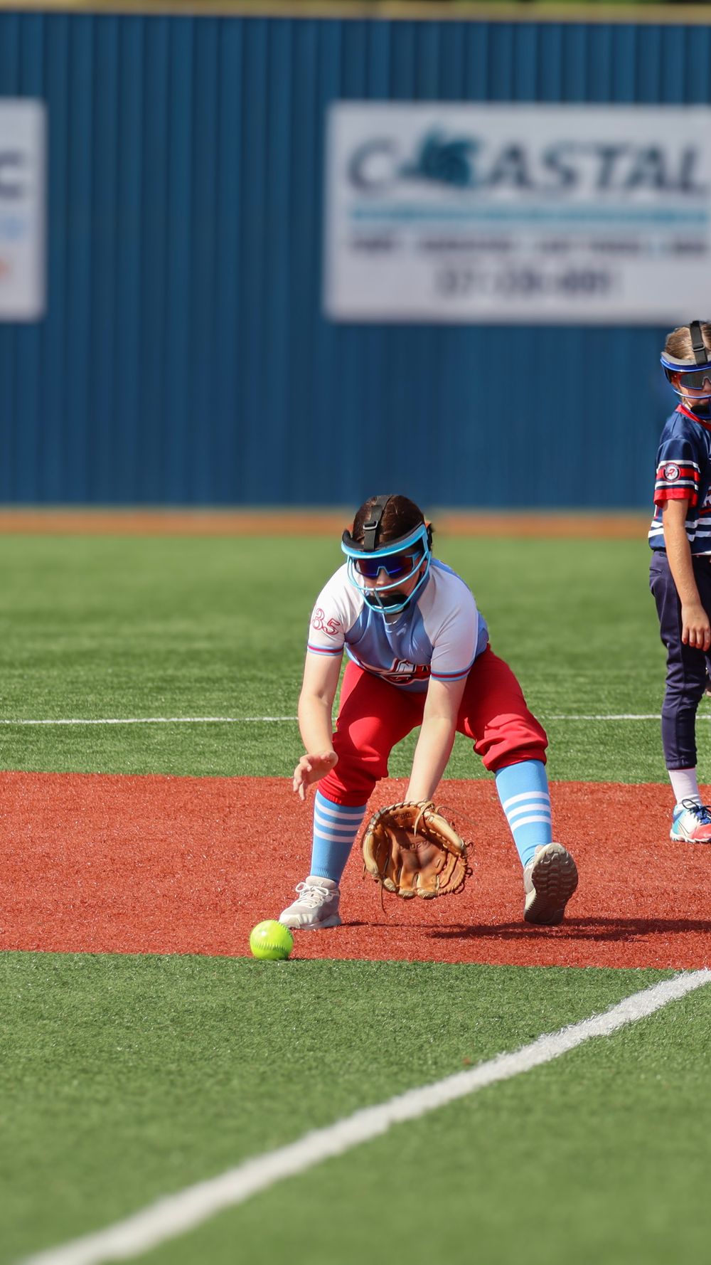 Softball infielder in red pants and protective mask fields ground ball during game.