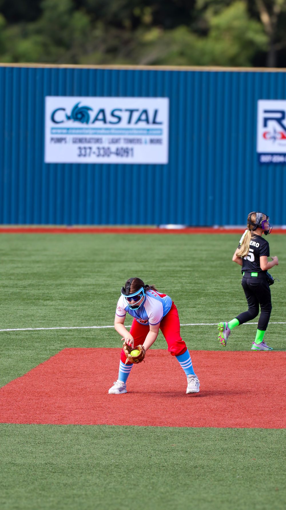 Player fielding ground ball on red turf infield with outfield fence visible.