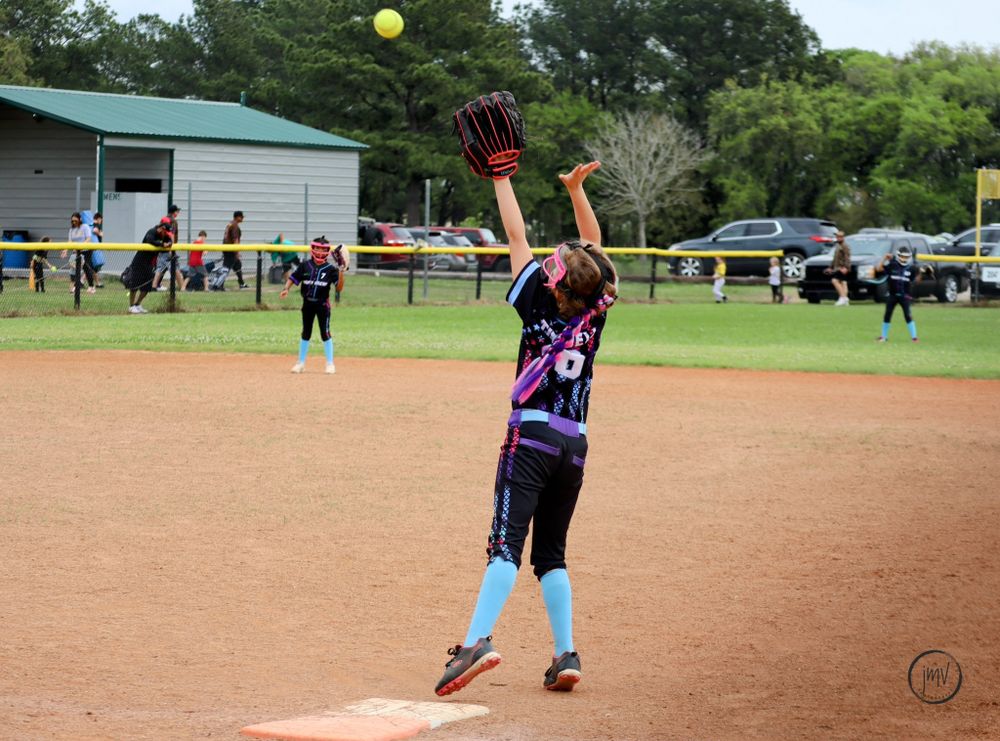 Yellow softball flying through air during game with players on dirt field.