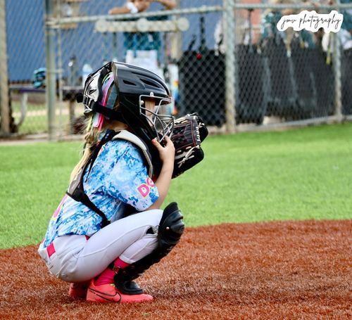 Softball catcher in tie-dye uniform and pink cleats crouches behind home plate.