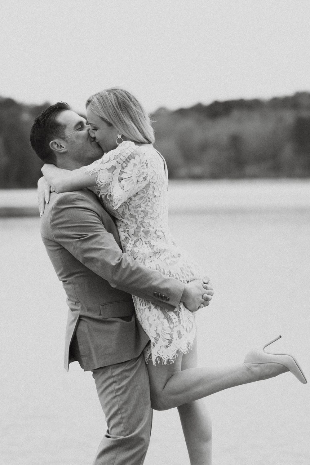 Black and white photo of an intimate embrace between a couple in formal attire by a lake.