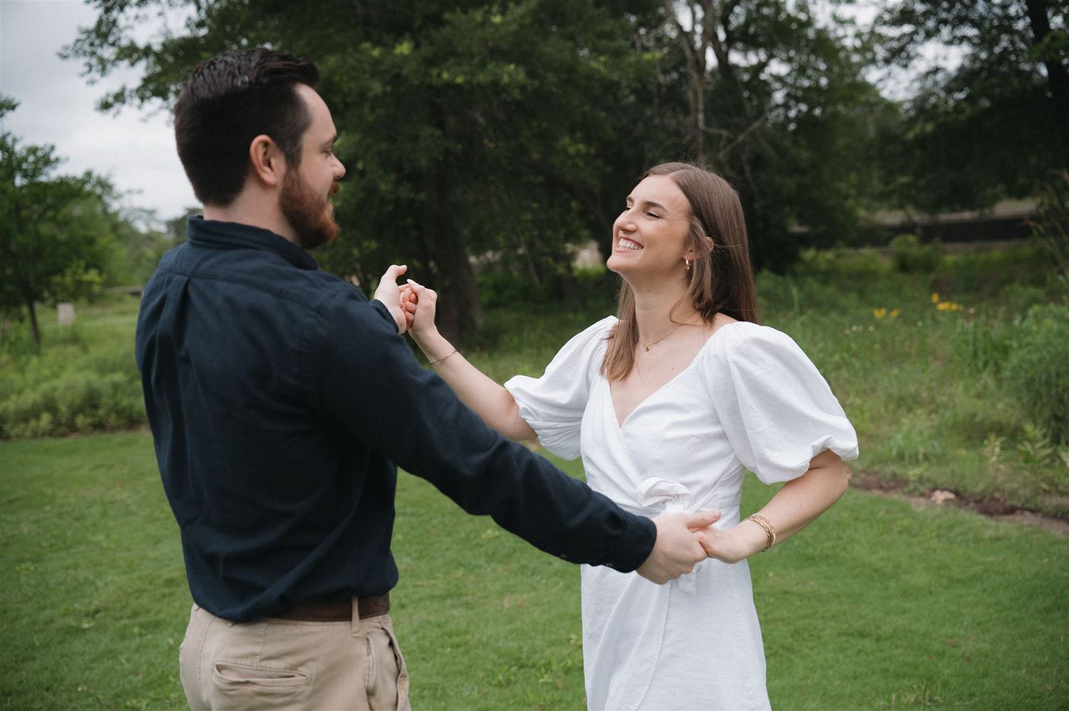 Two people in blue and white attire share a joyful moment dancing together on a green lawn in nature.