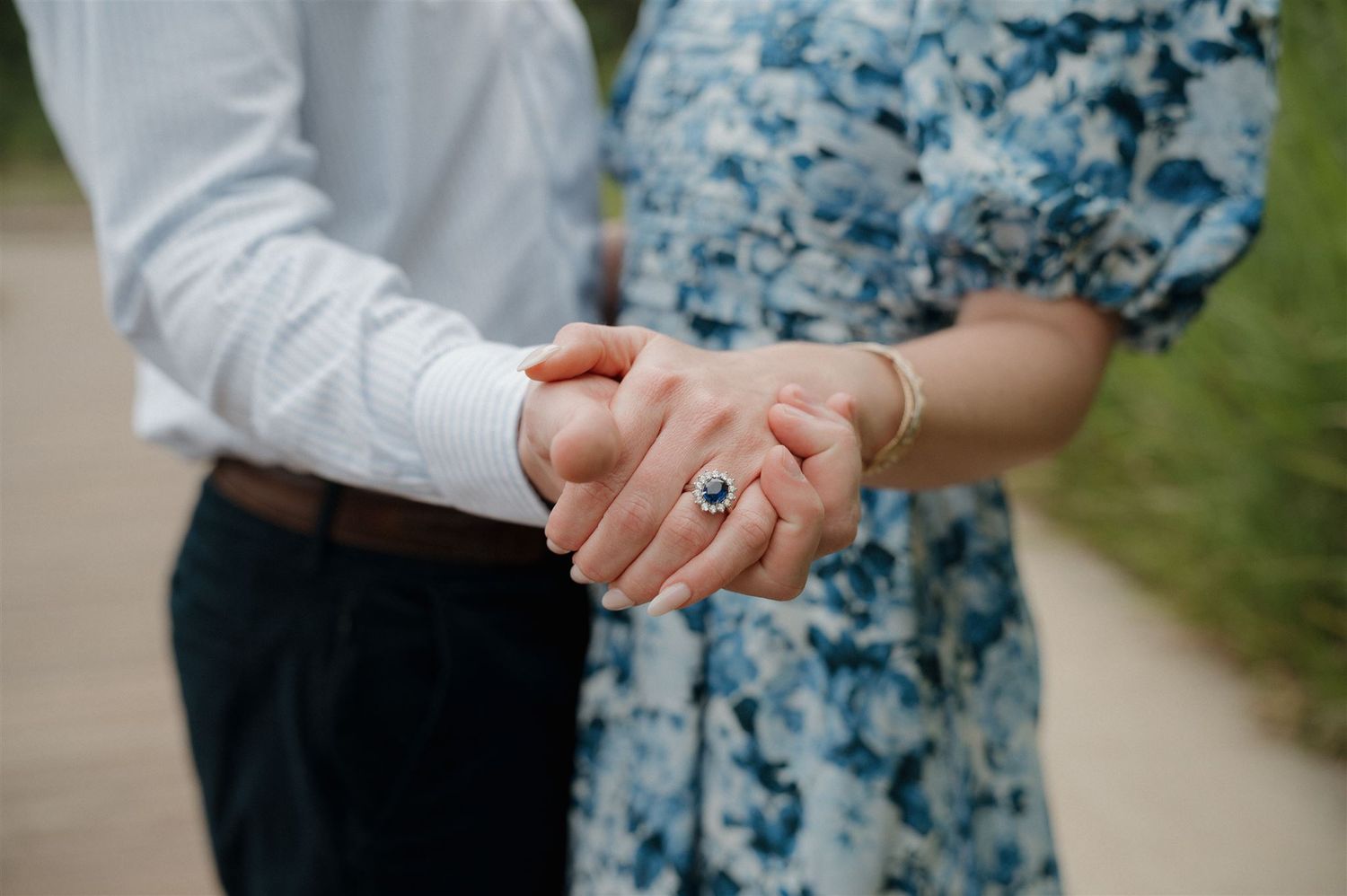 Close-up of two hands clasped together with an engagement ring, showing a romantic moment on a path outdoors.