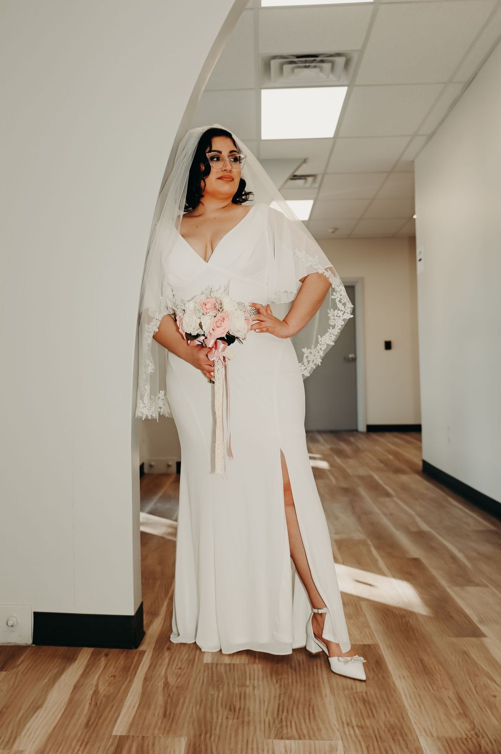 Bride in white wedding dress posing in indoor hallway with wooden floors and white walls.