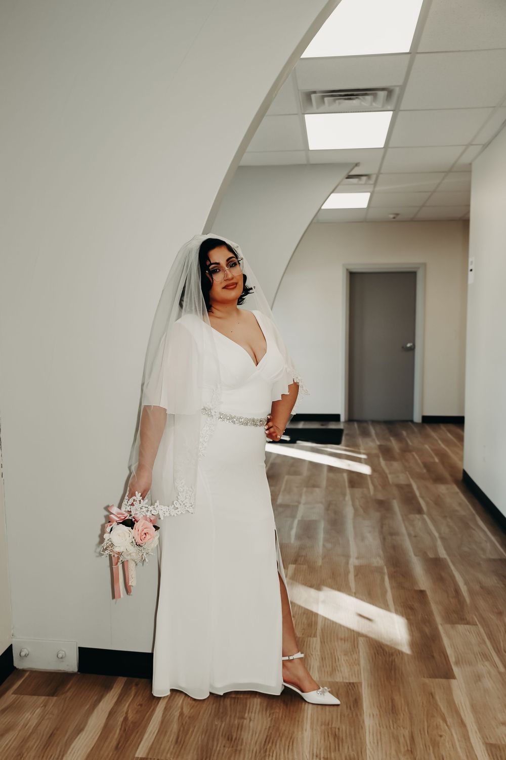 Bride in flowing white wedding gown standing in modern hallway with hardwood flooring.