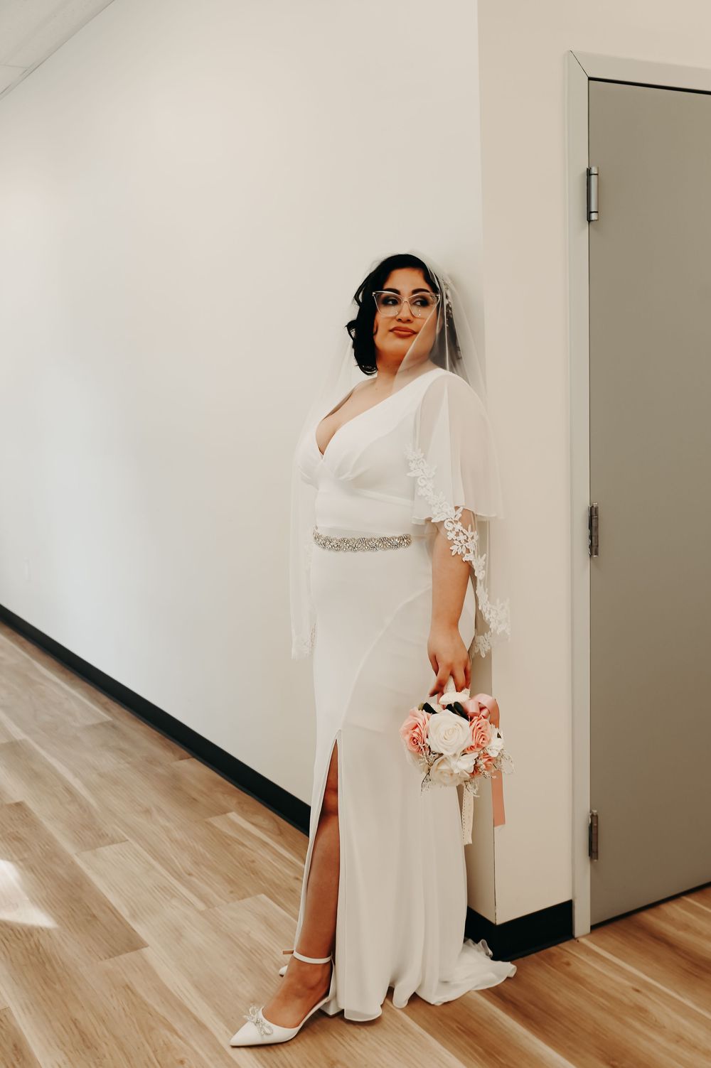Bride in elegant white wedding dress posing in modern indoor setting with wooden floors.