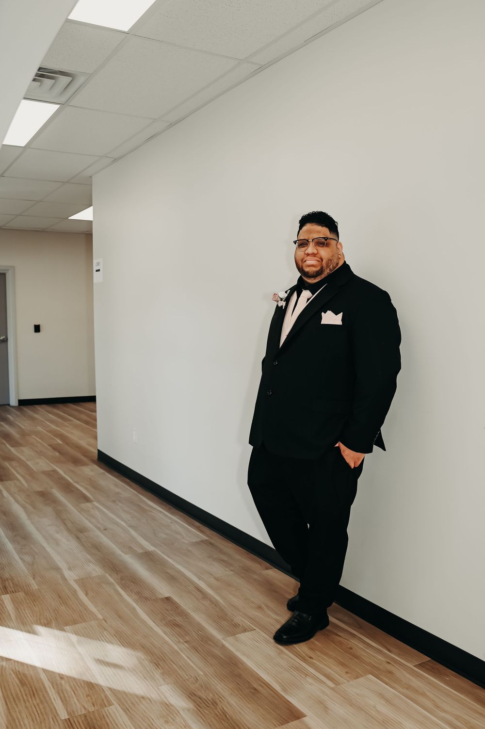 A person in a black suit poses against a white wall in a hallway.