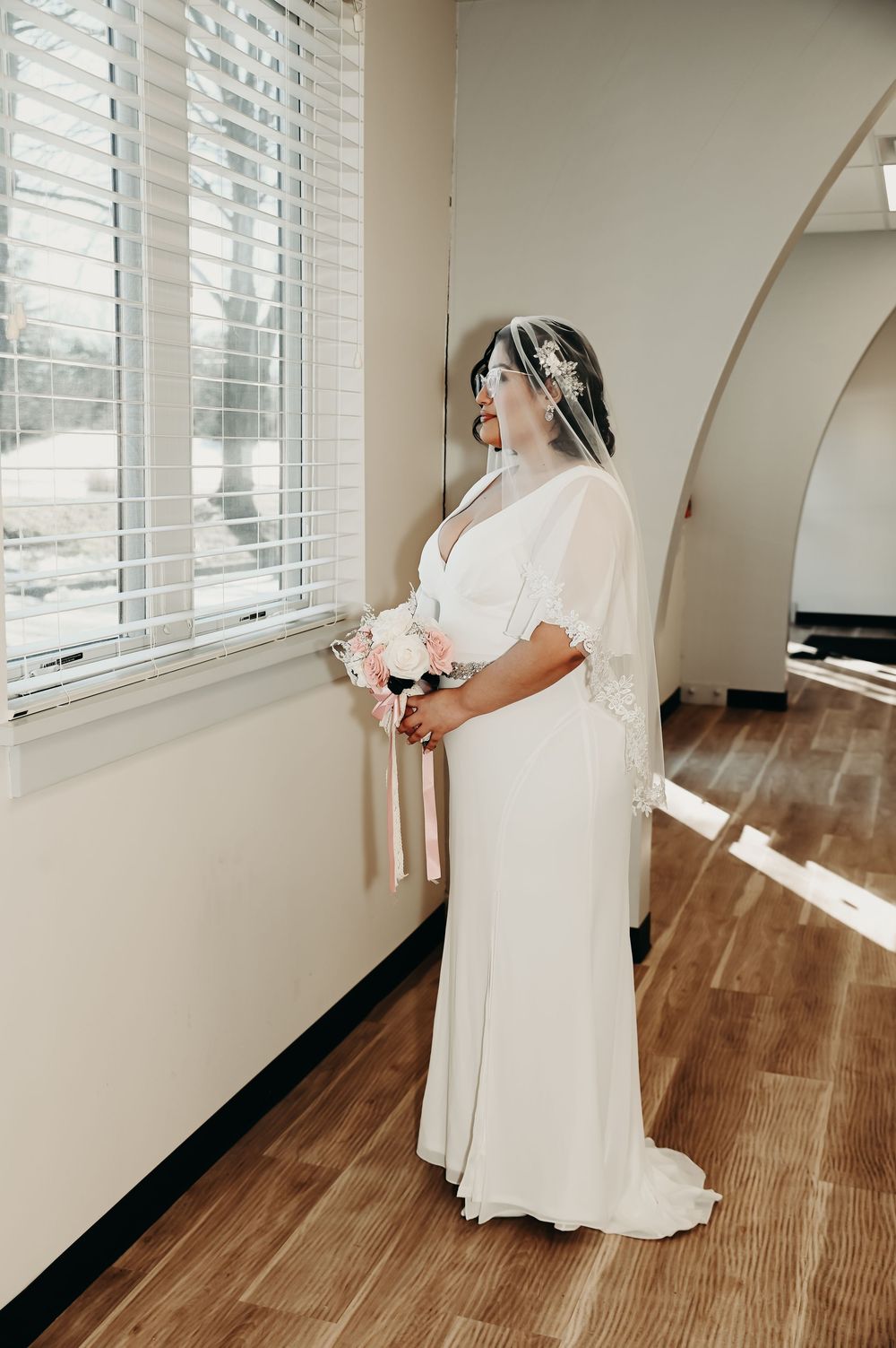 Bride in flowing white dress poses by a window with natural lighting.