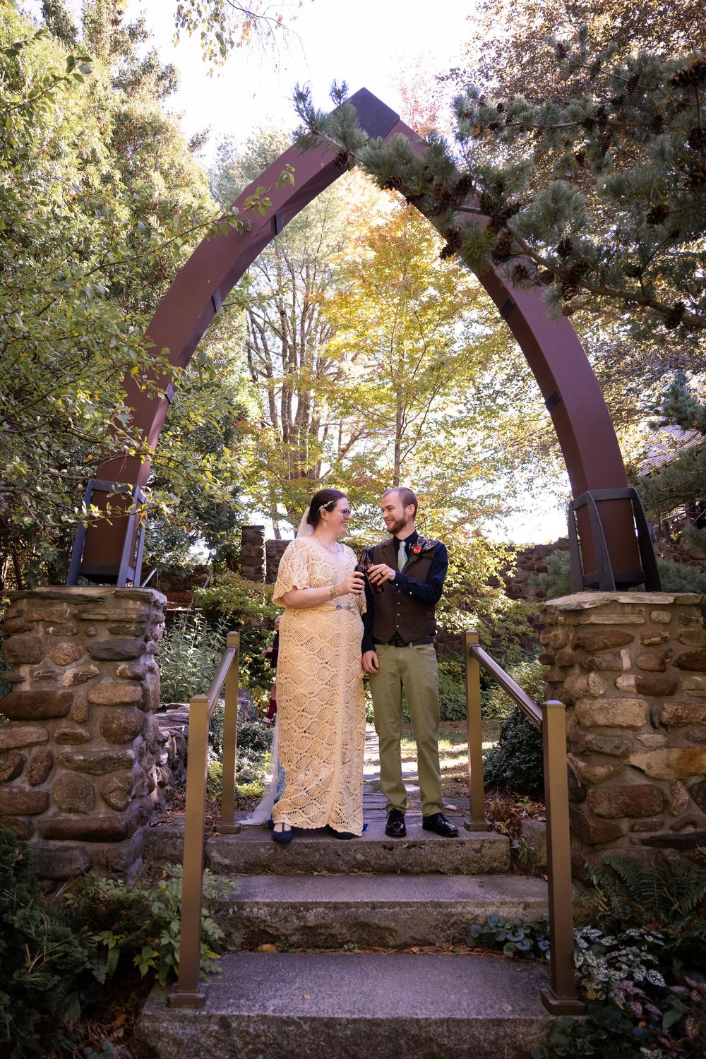 A couple poses beneath a rustic metal archway surrounded by autumn foliage and stone pillars.