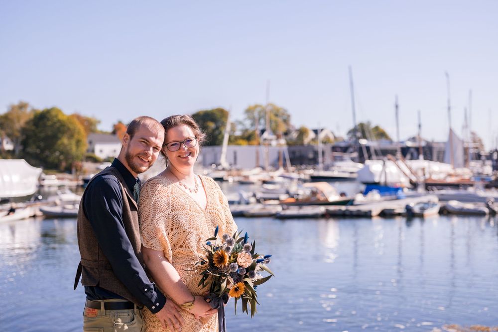 Couple embraces near a marina with boats and autumn trees in the background during sunset.