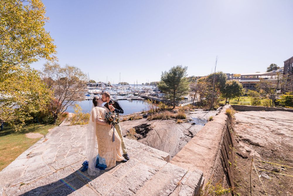Stone steps lead down a rocky coastal path with autumn trees and boats in the marina visible in the background.