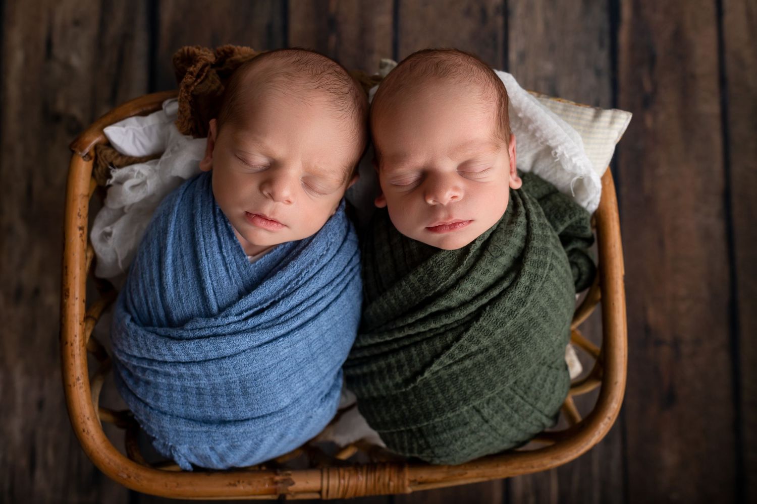 Twin newborns sleeping in wooden basket wrapped in blue and green swaddles.
