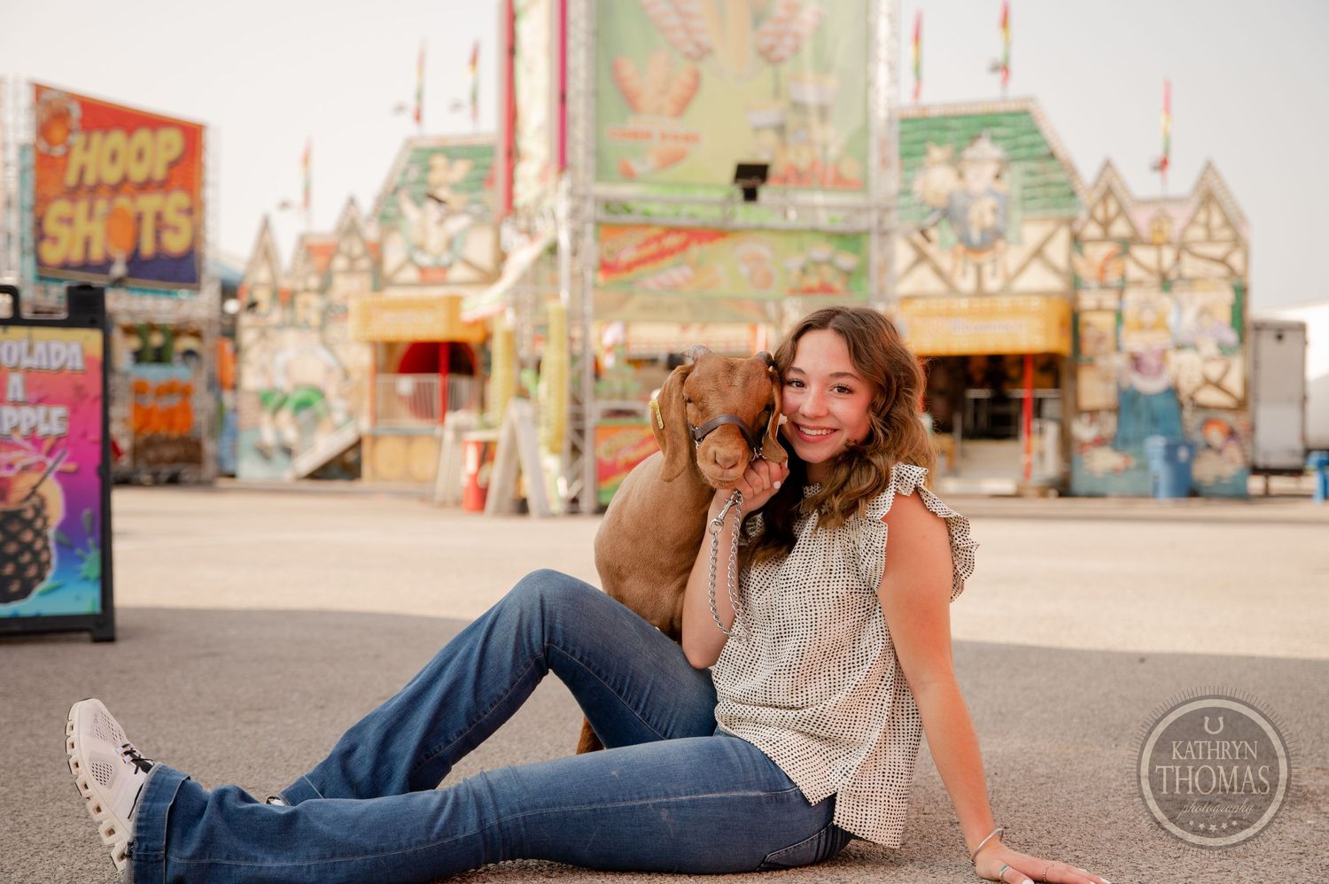 Tan goat rests with companion at carnival fairgrounds with colorful stands in background.