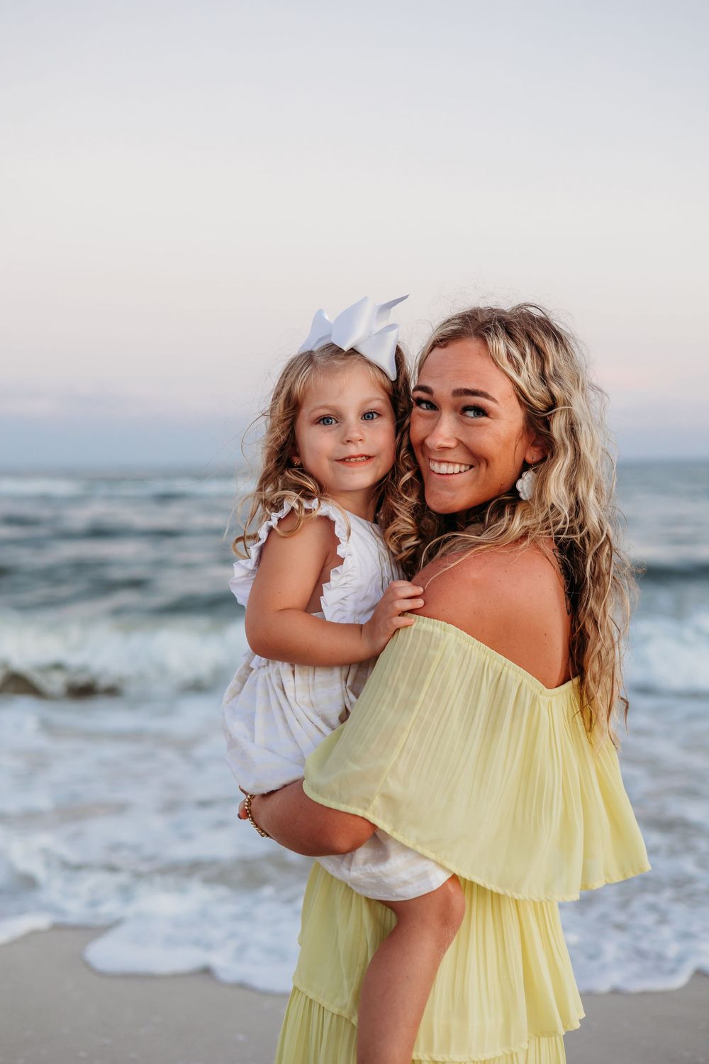 Mother and daughter in yellow and white dresses sharing tender moments by the ocean.