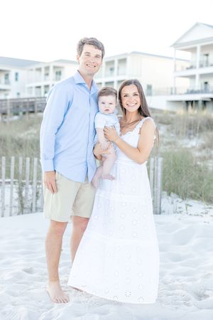 Happy young family with baby posing on sandy beach, man in blue shirt and woman in white dress.