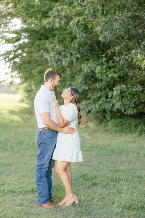 A romantic couple embraces in a sunlit meadow with green trees in the background.